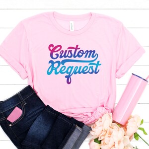 May include: Pink t-shirt with the text "Custom Request" in a colorful gradient font. The shirt is displayed on a white wooden background with blue jeans, a pink tumbler, and pink flowers.
