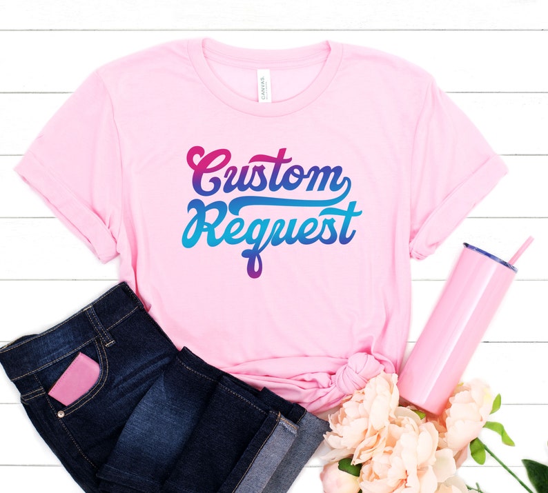 May include: Pink t-shirt with the text "Custom Request" in a colorful gradient font. The shirt is displayed on a white wooden background with blue jeans, a pink tumbler, and pink flowers.
