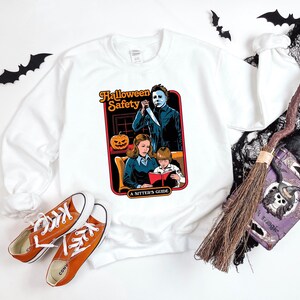 May include: White sweatshirt featuring a graphic of a vintage Halloween safety guide. The guide depicts a woman, a child, and a man holding a knife. The text reads "Halloween Safety A Sitter's Guide".