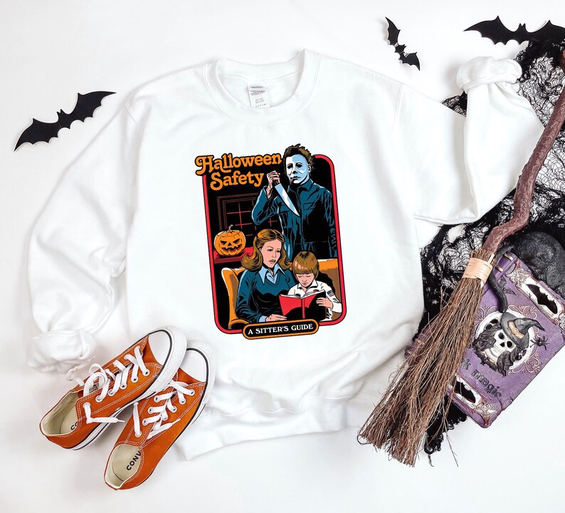 May include: White sweatshirt featuring a graphic of a vintage Halloween safety guide. The guide depicts a woman, a child, and a man holding a knife. The text reads "Halloween Safety A Sitter's Guide".