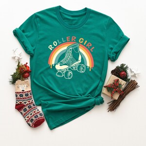 Peut inclure: Un t-shirt vert avec le texte "Roller Girl" dans un design arc-en-ciel r&eacute;tro. Le design pr&eacute;sente un patin &agrave; roulettes vintage avec des roues.