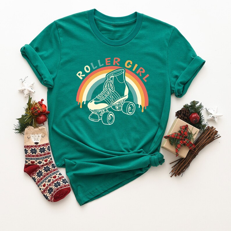 Peut inclure: Un t-shirt vert avec le texte "Roller Girl" dans un design arc-en-ciel r&eacute;tro. Le design pr&eacute;sente un patin &agrave; roulettes vintage avec des roues.