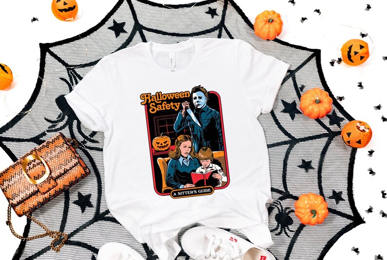 May include: White t-shirt with a graphic of a horror movie character holding a knife, with the text "Halloween Safety A Sitter's Guide" printed on the shirt.