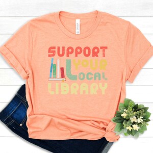May include: A peach-colored t-shirt with the text "SUPPORT YOUR LOCAL LIBRARY" in a retro-style font. The text is in various colors, including red, yellow, green, and blue. A stack of books is depicted next to the text.