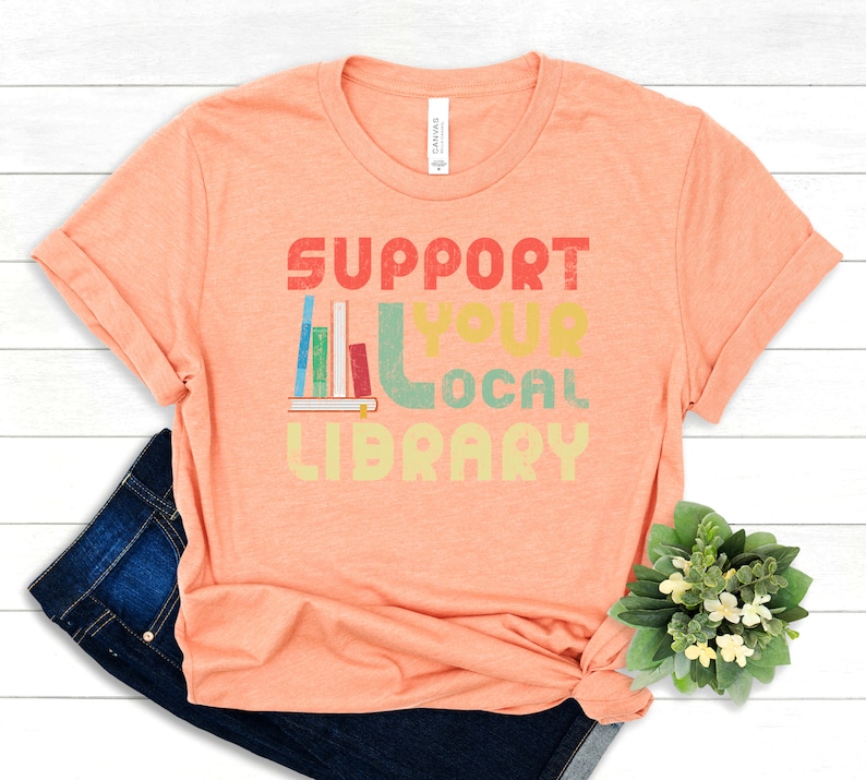 May include: A peach-colored t-shirt with the text "SUPPORT YOUR LOCAL LIBRARY" in a retro-style font. The text is in various colors, including red, yellow, green, and blue. A stack of books is depicted next to the text.