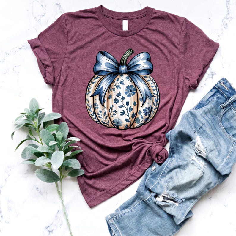K&ouml;nnte beinhalten: Burgunderfarbenes T-Shirt mit einem floralen K&uuml;rbisdesign und einer blauen Schleife. Der K&uuml;rbis hat einen cremefarbenen Hintergrund mit blauen floralen Akzenten. Das Shirt wird mit blauen Jeans kombiniert. Ein Zweig gr&uuml;ner Bl&auml;tter befindet sich links.