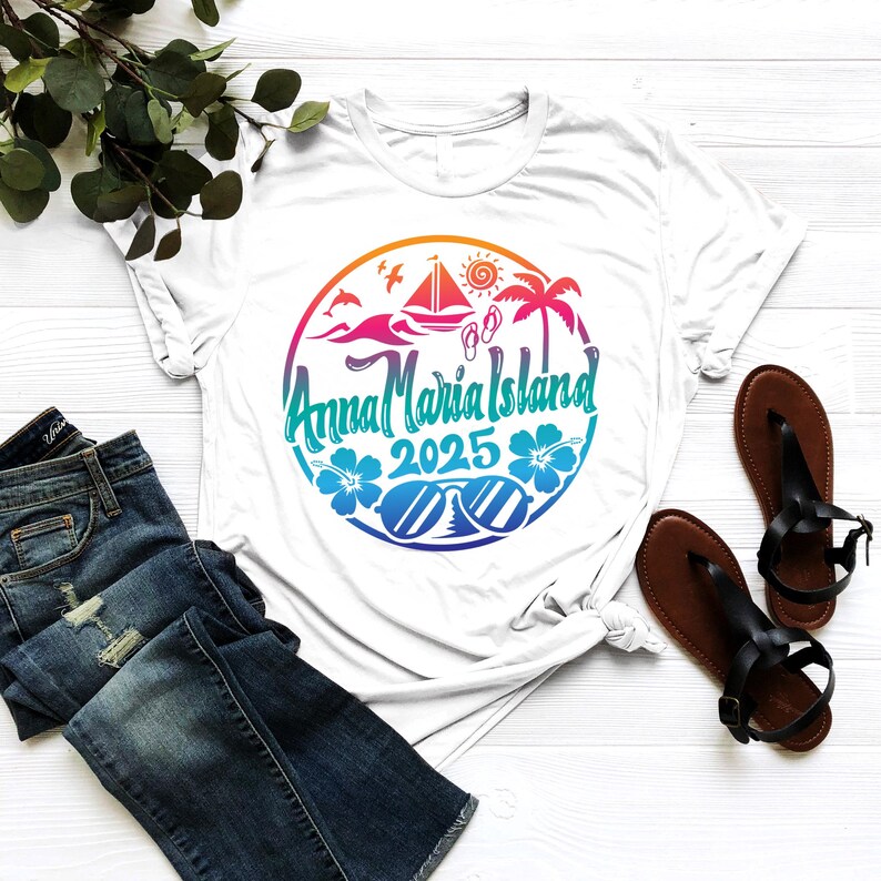 Puede incluir: Camiseta blanca con un dise&ntilde;o gr&aacute;fico colorido que presenta un c&iacute;rculo con el texto "Anna Maria Island 2025" en su interior. El gr&aacute;fico tambi&eacute;n incluye un velero, palmeras, gafas de sol y flores.