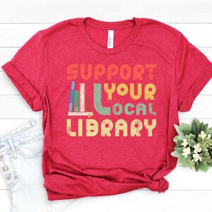 May include: Red t-shirt with the text "Support Your Local Library" in a retro font with a stack of colorful books.