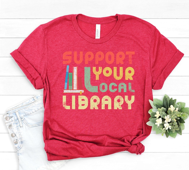 May include: Red t-shirt with the text "Support Your Local Library" in a retro font with a stack of colorful books.