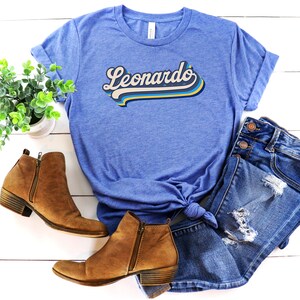 Pode incluir: Uma camiseta azul com um gr&aacute;fico de estilo retr&ocirc; que diz "Leonardo" em uma fonte amarela, azul e branca. A camiseta &eacute; combinada com jeans azul e botas de tornozelo marrons.