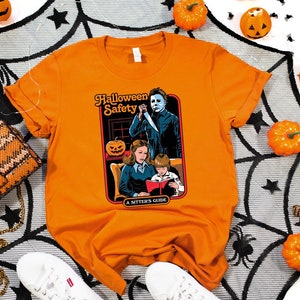 May include: Orange t-shirt with a graphic of a horror movie character holding a knife and a family reading a book. The graphic says "Halloween Safety A Sitter's Guide".