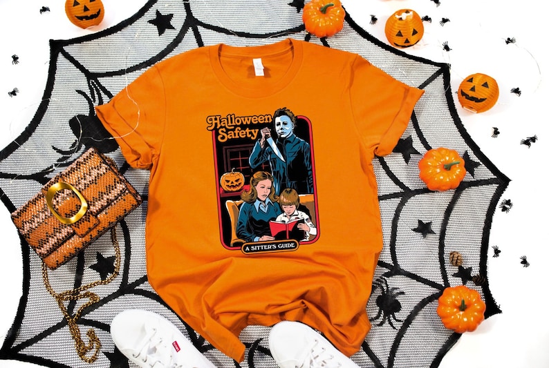 May include: Orange t-shirt with a graphic of a horror movie character holding a knife and a family reading a book. The graphic says "Halloween Safety A Sitter's Guide".