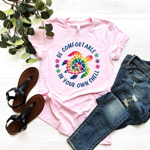 Może przedstawiać: Jasnor&oacute;żowa koszulka z nadrukiem ż&oacute;łwia tie-dye i napisem &bdquo;BE COMFORTABLE IN YOUR OWN SHELL&rdquo;. Na zdjęciu znajdują się r&oacute;wnież niebieskie dżinsy i czarne sandały.