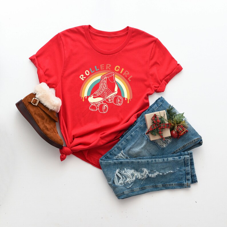Peut inclure: T-shirt rouge avec un graphique arc-en-ciel et le texte "Roller Girl". Le graphique repr&eacute;sente un patin &agrave; roulettes avec un contour blanc. Le t-shirt est pos&eacute; sur une surface blanche avec une paire de bottes brunes, un jean bleu et une petite bo&icirc;te cadeau.