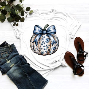 K&ouml;nnte beinhalten: Wei&szlig;es T-Shirt mit einem K&uuml;rbisdesign, einer blauen Schleife und einem floralen Muster. Der K&uuml;rbis hat eine hellbraune Basis mit blauen floralen Akzenten. Das T-Shirt wird mit blauen Jeans und braunen Sandalen kombiniert.