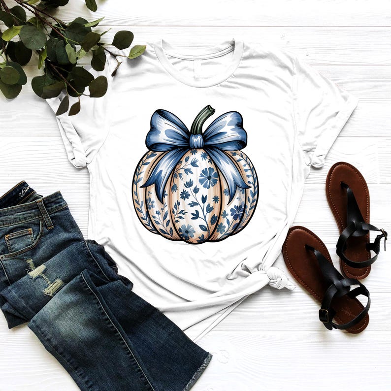 K&ouml;nnte beinhalten: Wei&szlig;es T-Shirt mit einem K&uuml;rbisdesign, einer blauen Schleife und einem floralen Muster. Der K&uuml;rbis hat eine hellbraune Basis mit blauen floralen Akzenten. Das T-Shirt wird mit blauen Jeans und braunen Sandalen kombiniert.