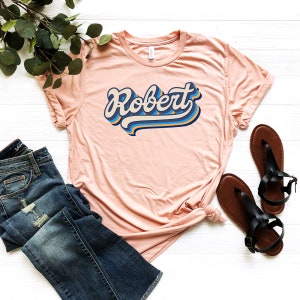 Pode incluir: Uma camiseta rosa claro com o nome "Robert" estampado em uma fonte retr&ocirc; e colorida. A camiseta est&aacute; colocada sobre uma superf&iacute;cie branca com um par de sand&aacute;lias marrons e uma cal&ccedil;a jeans azul.