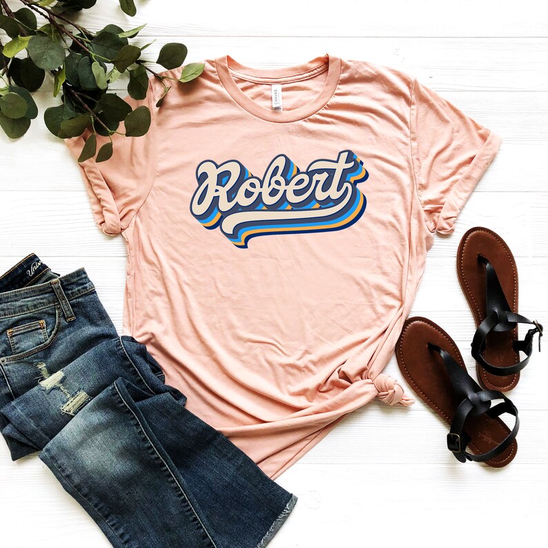 Pode incluir: Uma camiseta rosa claro com o nome "Robert" estampado em uma fonte retr&ocirc; e colorida. A camiseta est&aacute; colocada sobre uma superf&iacute;cie branca com um par de sand&aacute;lias marrons e uma cal&ccedil;a jeans azul.