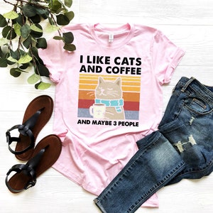 May include: Pink t-shirt featuring a retro-style graphic of a cat holding a cup of coffee. The text on the shirt reads "I like cats and coffee and maybe 3 people".