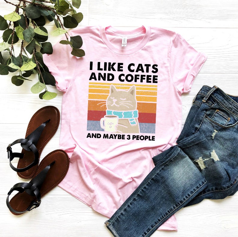 May include: Pink t-shirt featuring a retro-style graphic of a cat holding a cup of coffee. The text on the shirt reads "I like cats and coffee and maybe 3 people".