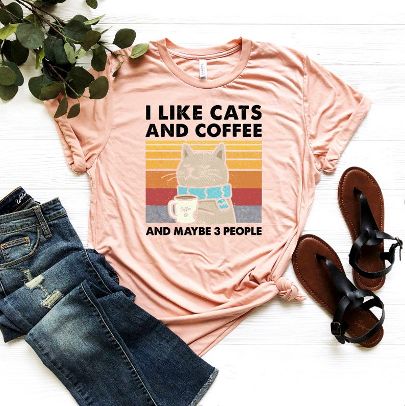 May include: A peach-coloured t-shirt with the text "I like cats and coffee and maybe 3 people" in black. A retro-style illustration of a cat wearing a blue scarf and holding a cup of coffee is above the text.