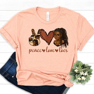 May include: Peach colored t-shirt with a peace sign, heart, and a woman with locs. The text reads "peace love locs".
