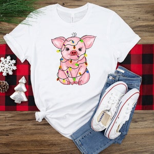 May include: White t-shirt with a pink pig wearing colorful Christmas lights. The pig is sitting down with its legs crossed. The t-shirt is on a red and black checkered surface.
