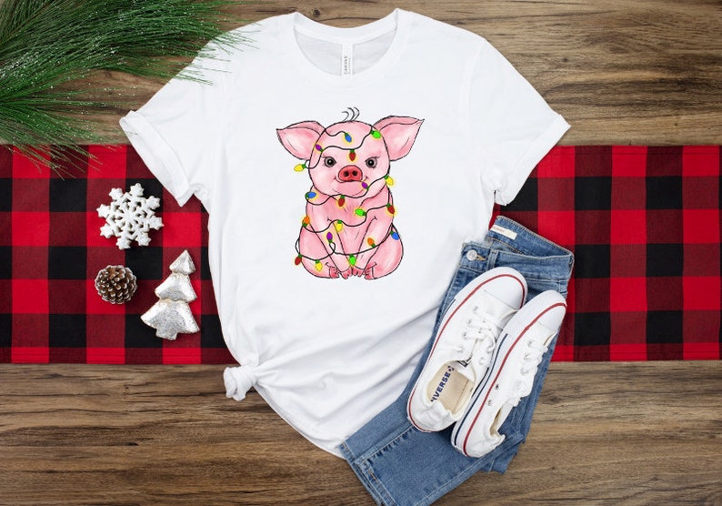 May include: White t-shirt with a pink pig wearing colorful Christmas lights. The pig is sitting down with its legs crossed. The t-shirt is on a red and black checkered surface.