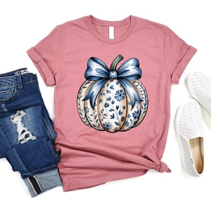 K&ouml;nnte beinhalten: Staubrosa T-Shirt mit einem K&uuml;rbis-Design. Der K&uuml;rbis hat eine blaue Schleife und blaue und cremefarbene Blumenmuster. Das Shirt wird mit blauen Jeans und wei&szlig;en Slip-on-Schuhen kombiniert. Herbstliches Thema.