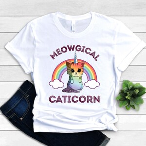 May include: White t-shirt with a rainbow and a caticorn illustration. The text "MEOWGICAL CATICORN" is printed on the shirt.