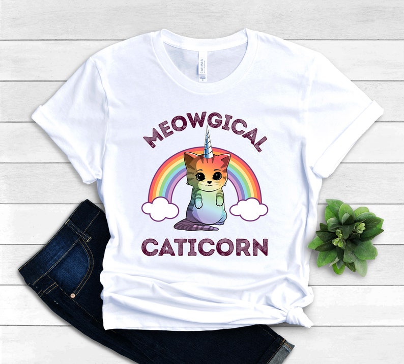 May include: White t-shirt with a rainbow and a caticorn illustration. The text "MEOWGICAL CATICORN" is printed on the shirt.