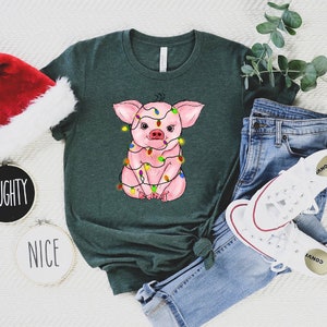 May include: A green t-shirt with a pink pig wearing Christmas lights. The pig is sitting down and looking at the camera. The t-shirt is on a white surface with a Santa hat, a pair of jeans, and a pair of white sneakers.