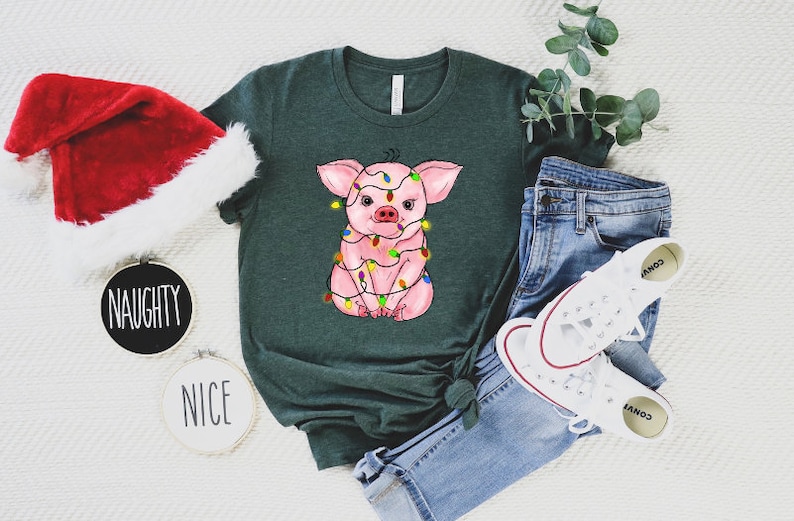 May include: A green t-shirt with a pink pig wearing Christmas lights. The pig is sitting down and looking at the camera. The t-shirt is on a white surface with a Santa hat, a pair of jeans, and a pair of white sneakers.