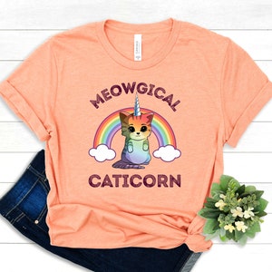 May include: A peach-colored t-shirt with a rainbow and a cartoon cat with a unicorn horn. The text "MEOWGICAL CATICORN" is printed on the shirt.
