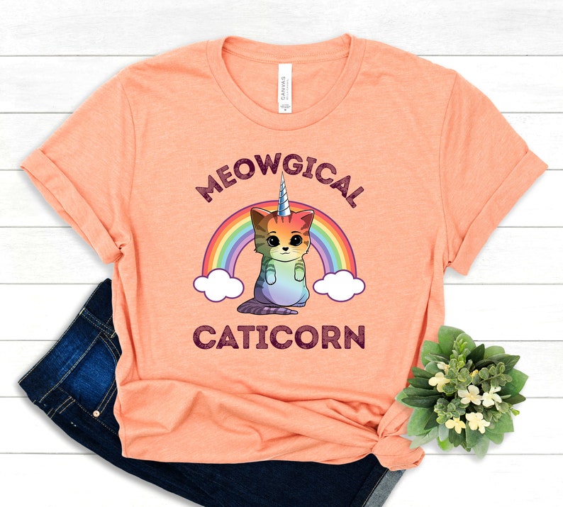 May include: A peach-colored t-shirt with a rainbow and a cartoon cat with a unicorn horn. The text "MEOWGICAL CATICORN" is printed on the shirt.