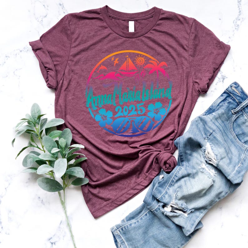 Puede incluir: Una camiseta de color granate con un dise&ntilde;o gr&aacute;fico colorido de una escena de isla tropical. El dise&ntilde;o incluye un velero, palmeras y el texto "Annual Island 2025".