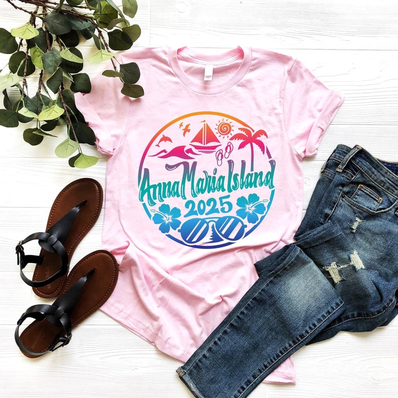 Puede incluir: Camiseta rosa con un dise&ntilde;o gr&aacute;fico colorido de una escena de playa con un velero, palmeras, gafas de sol y el texto "Anna Maria Island 2025".