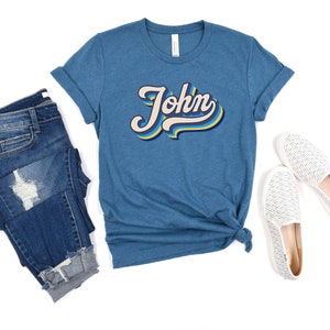 Pode incluir: Uma camiseta azul turquesa com um gr&aacute;fico de estilo retr&ocirc; do nome "John" em amarelo, azul e branco.