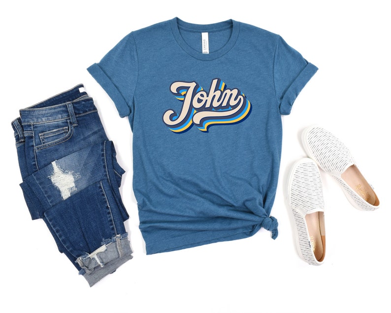 Pode incluir: Uma camiseta azul turquesa com um gr&aacute;fico de estilo retr&ocirc; do nome "John" em amarelo, azul e branco.