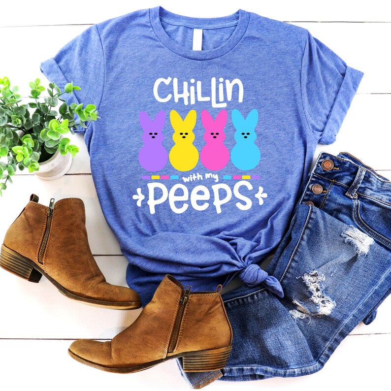 May include: A blue t-shirt with the text "Chillin with my Peeps" and three colorful marshmallow Peeps. The shirt is laid out on a white wooden surface with a pair of brown ankle boots and blue jeans.