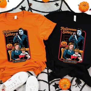 May include: Two t-shirts with a Halloween safety graphic featuring a scary character holding a knife. The graphic is a parody of a children's book cover. The text on the graphic reads "Halloween Safety A Sitter's Guide". One shirt is orange and the other is black.