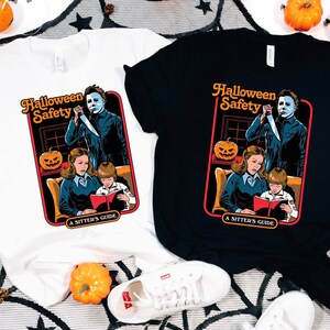 May include: Two t-shirts with a Halloween Safety graphic. The graphic features a woman reading a book to a child while a man with a knife stands behind them. The text on the graphic says "Halloween Safety A Sitter's Guide". One shirt is white and the other is black.