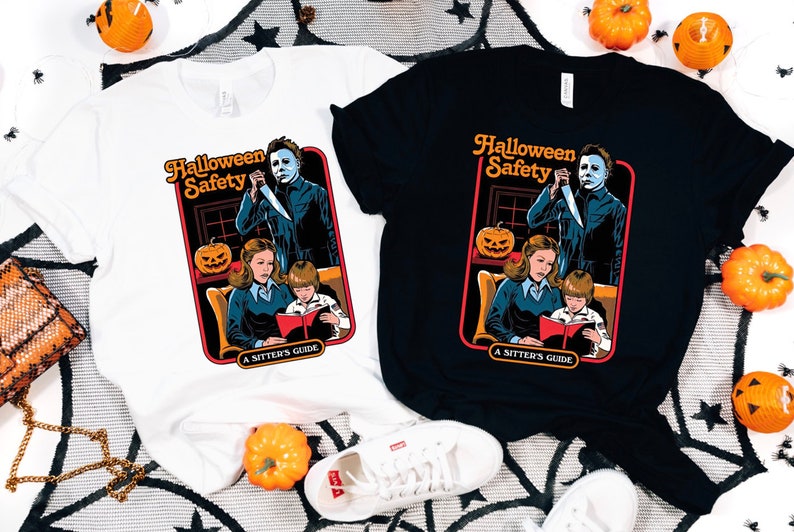 May include: Two t-shirts with a Halloween Safety graphic. The graphic features a woman reading a book to a child while a man with a knife stands behind them. The text on the graphic says "Halloween Safety A Sitter's Guide". One shirt is white and the other is black.
