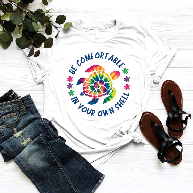 Może przedstawiać: Biała koszulka z nadrukiem ż&oacute;łwia tie-dye i napisem "BE COMFORTABLE IN YOUR OWN SHELL" w kolorze niebieskim. Koszulka jest połączona z niebieskimi dżinsami i czarnymi sandałami. Gałązka zielonych liści znajduje się w tle.