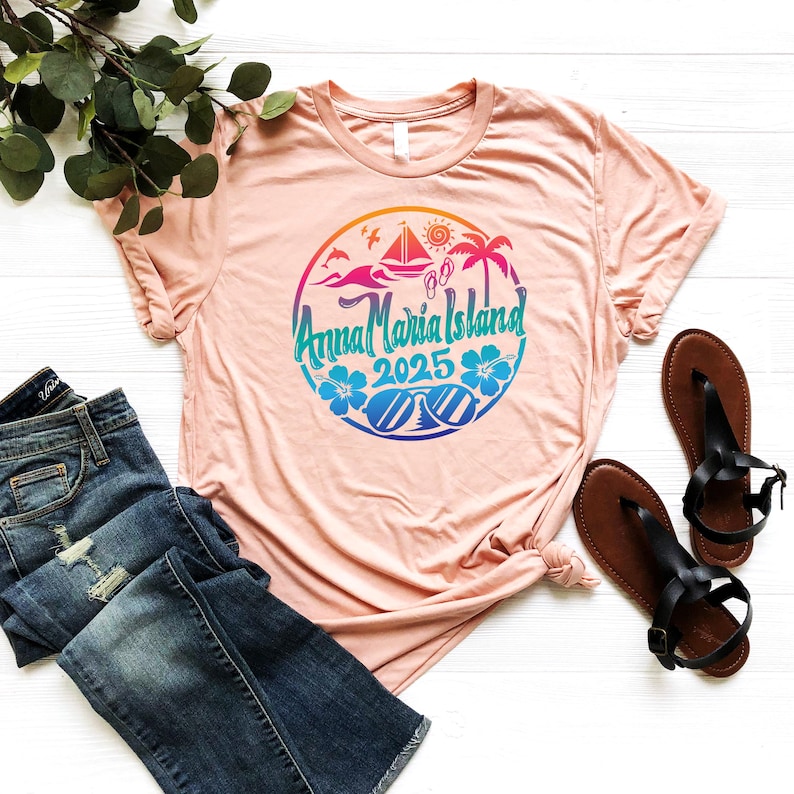 Puede incluir: Una camiseta rosa con un dise&ntilde;o gr&aacute;fico colorido que presenta un c&iacute;rculo con el texto "Anna Maria Island 2025" e im&aacute;genes con tem&aacute;tica de playa como un velero, palmeras, gafas de sol y chanclas.