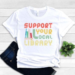 May include: White t-shirt with a vintage-style graphic that says "Support Your Local Library" with a stack of books and a bookend.