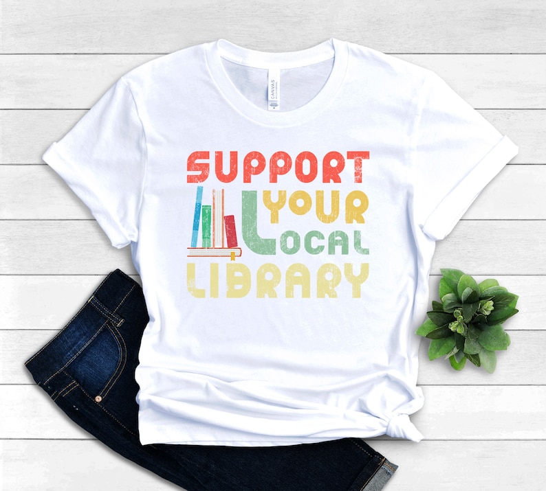 May include: White t-shirt with a vintage-style graphic that says "Support Your Local Library" with a stack of books and a bookend.