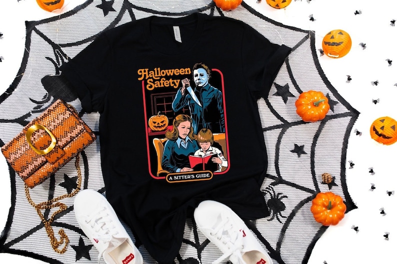 May include: Black t-shirt with a graphic of a horror movie-themed "Halloween Safety" guide. The guide features a woman reading a book to a child while a man with a knife stands behind them. The text on the guide reads "Halloween Safety, A Sitter's Guide."