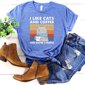 May include: A blue t-shirt featuring a vintage-style graphic of a cat wearing a scarf and holding a cup of coffee. The text on the shirt reads "I like cats and coffee and maybe 3 people".