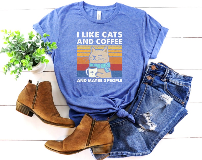 May include: A blue t-shirt featuring a vintage-style graphic of a cat wearing a scarf and holding a cup of coffee. The text on the shirt reads "I like cats and coffee and maybe 3 people".